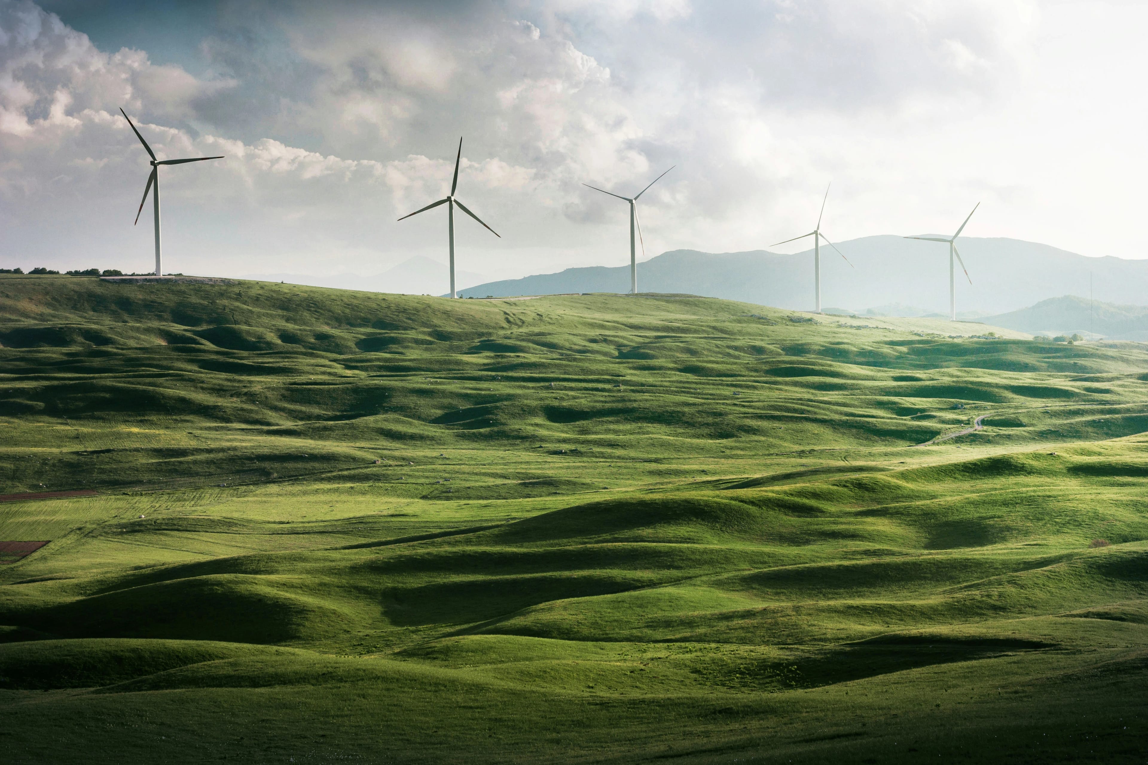 lush green hills with four wind turbine on top of them and big range mountains on the background